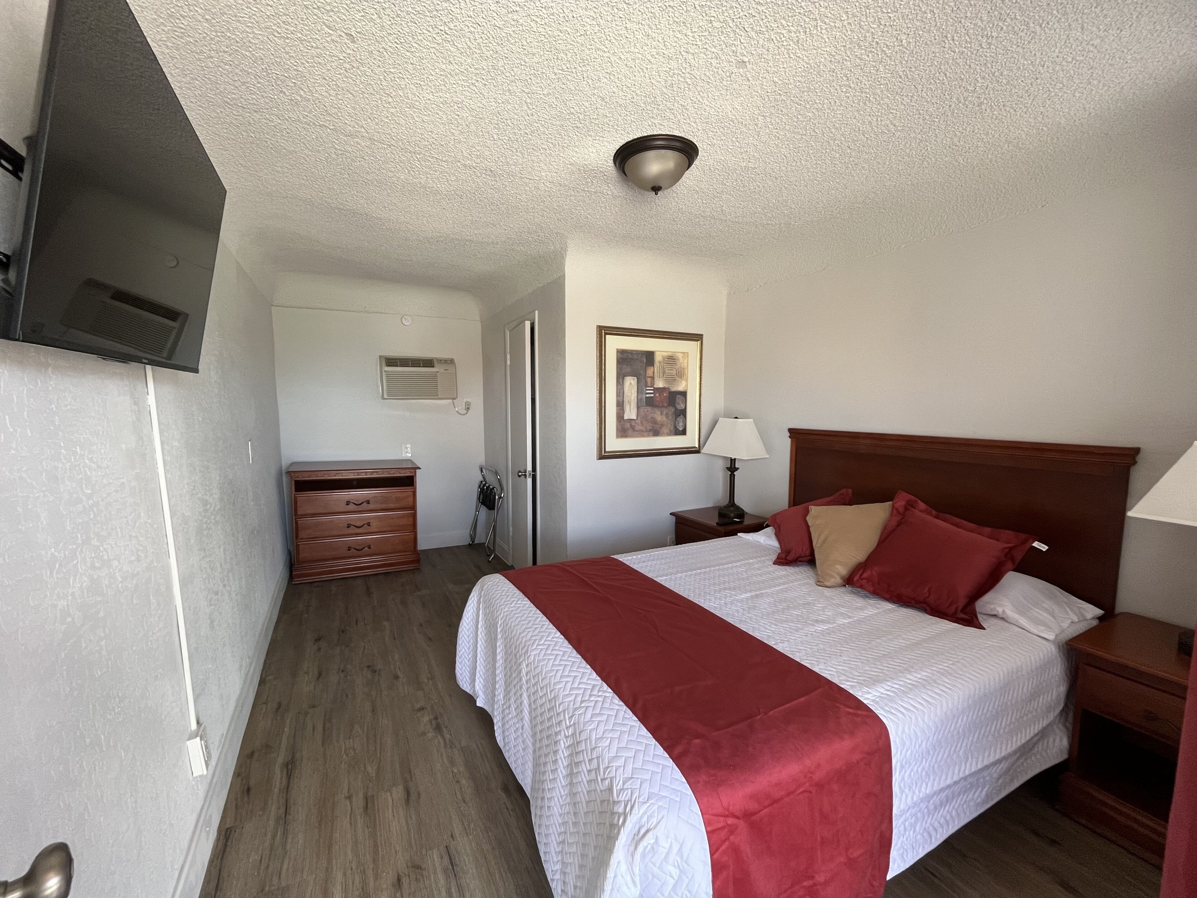 A hotel room with a bed, dresser, nightstand, lamp, air conditioning unit, and a ceiling vent.