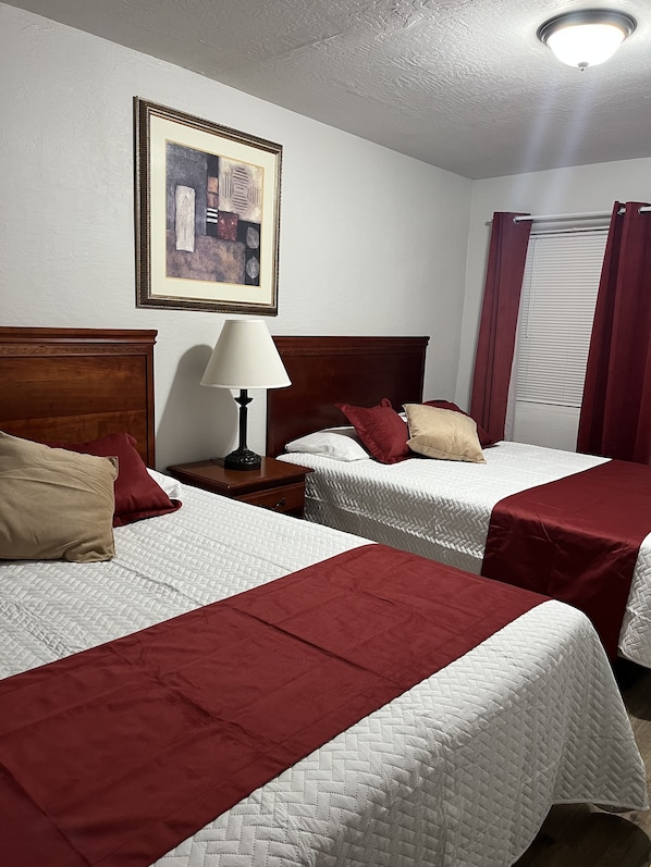 A hotel room with two beds, a nightstand with a lamp, a framed picture on the wall, and a window with curtains. - Arizona 9 Motor Hotel (Williams)