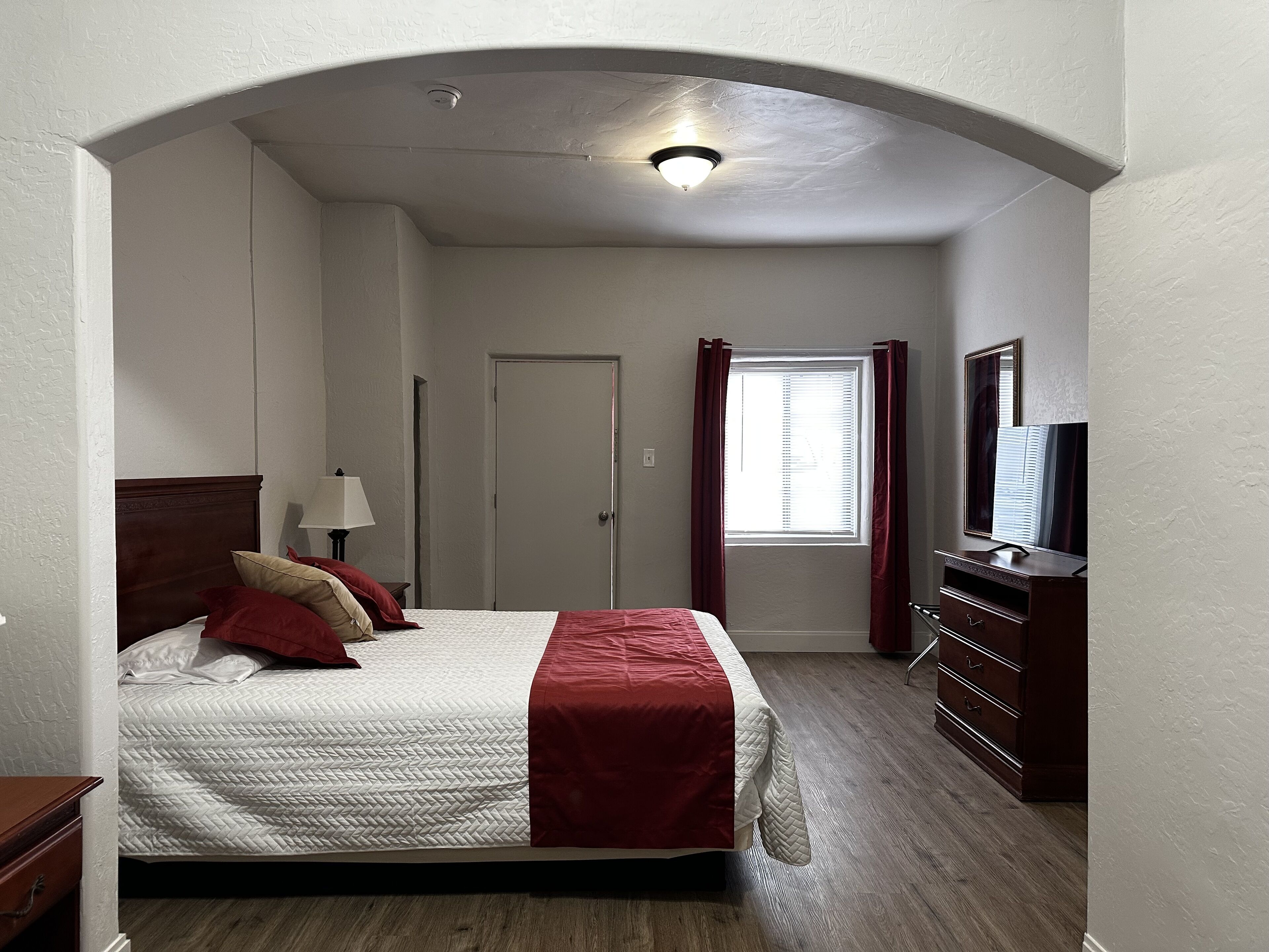 A bedroom with a bed, a dresser, a TV, a window with curtains, and a door.