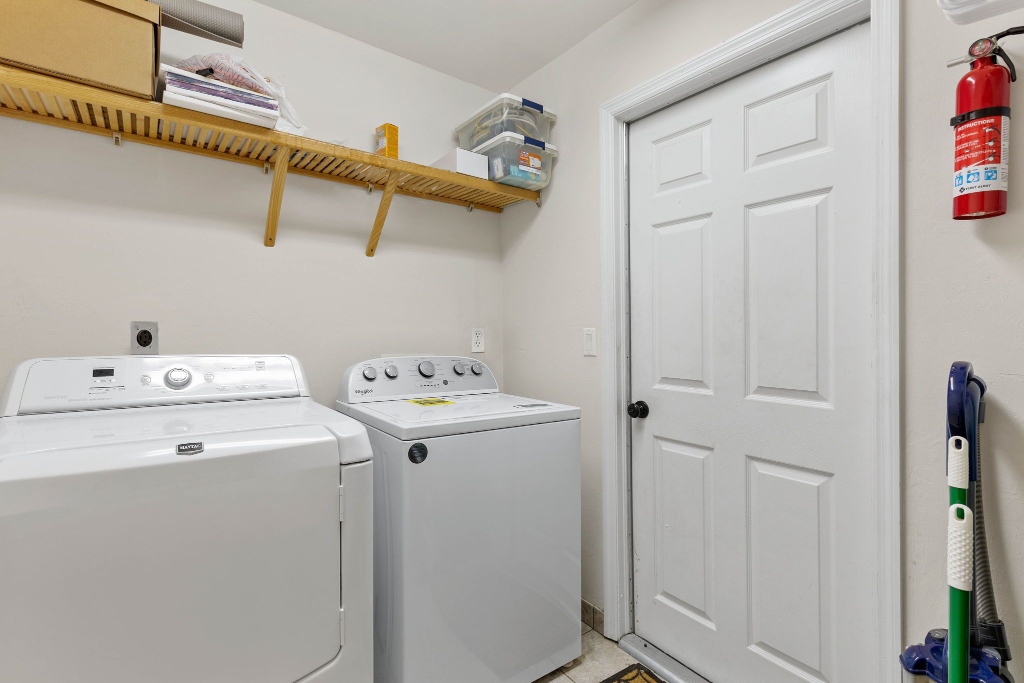A laundry area with two white washing machines, a shelf with storage boxes, a fire extinguisher, and a mop.