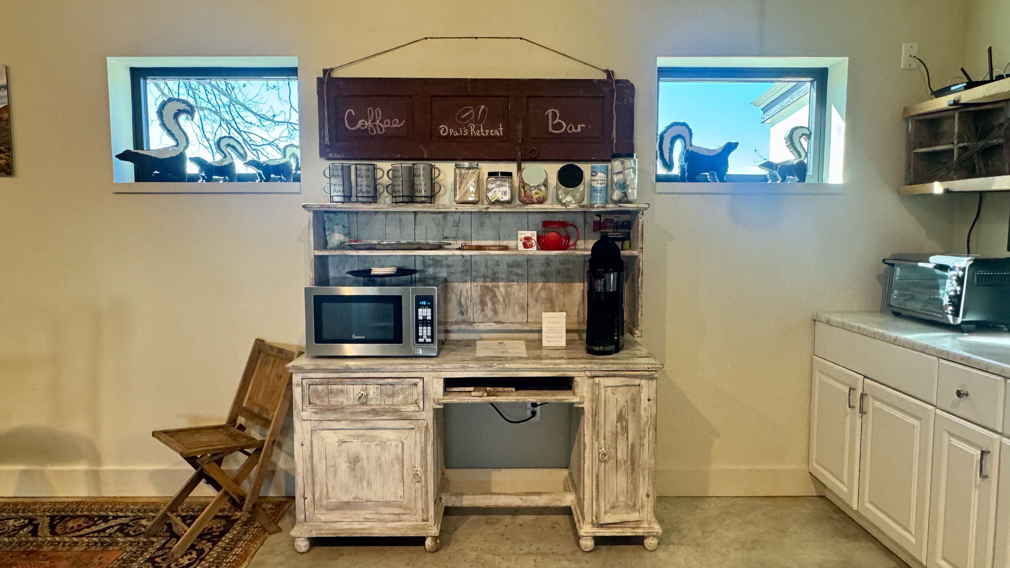 Una cocina con un escritorio de madera rústica, microondas, cafetera y tostadora. Hay dos ventanas con siluetas decorativas de ardillas.