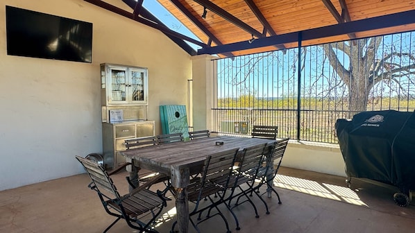 An outdoor patio with a wooden table and chairs, a grill, and a view of the countryside.