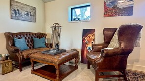 A living room with a brown leather sofa, a wooden coffee table, and a brown leather armchair. There are paintings on the walls and a window with a view of trees.