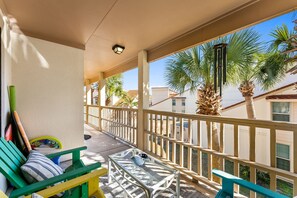 Un balcon avec une table en verre, des chaises vertes et une balustrade. On aperçoit des palmiers et un immeuble à l’arrière-plan.