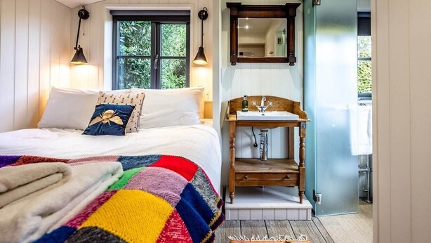 A bedroom with a bed, a colorful quilt, two wall-mounted lamps, a wooden bedside table with a sink, and a mirror. - The Little House, Orford (Orford)