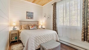 Un dormitorio con techo de madera, una cama con colcha blanca, dos lámparas de noche, una mesita de noche con lámpara, un cuadro enmarcado en la pared y una ventana con cortinas traslúcidas.