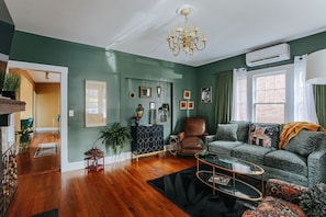 A living room with a green wall, wooden flooring, a brown leather armchair, a green sofa with patterned cushions, a glass coffee table, and a chandelier.