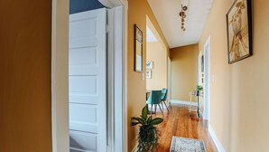 A hallway with a wooden floor, a white door, a dining area with chairs and a table, and a plant.