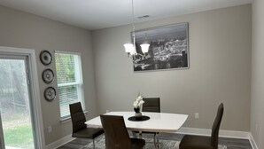 Un coin repas avec une table blanche, quatre chaises, un vase de fleurs, un lustre et un tableau représentant un paysage urbain accroché au mur.