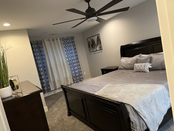 A bedroom with a bed, a nightstand, a desk, a ceiling fan, and patterned curtains.