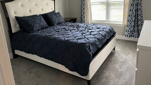 Un lit bien fait, avec une couette bleu foncé et des oreillers assortis, une tête de lit blanche et une fenêtre ornée de rideaux à motifs.