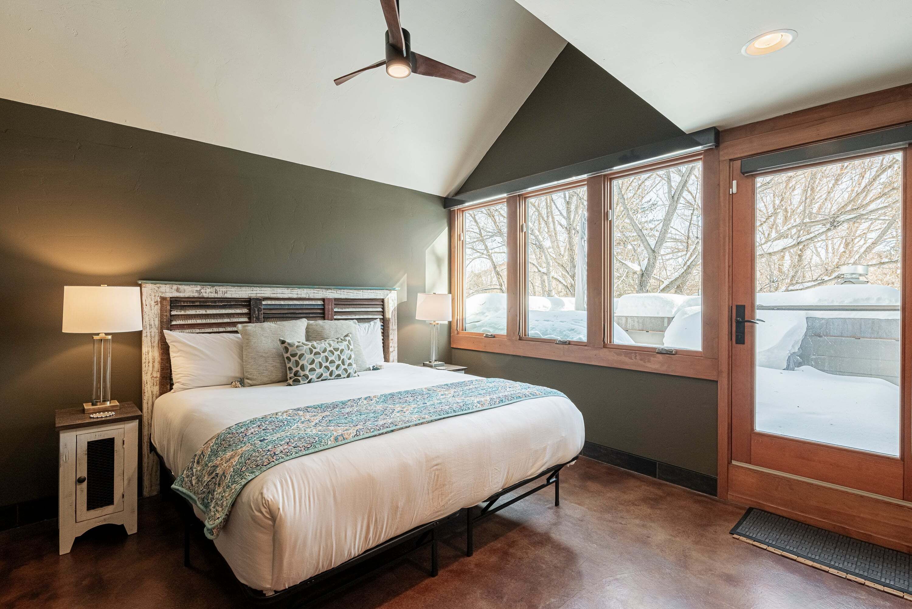 A bedroom with a bed, bedside lamps, a ceiling fan, and a large window with a view of a snowy landscape.