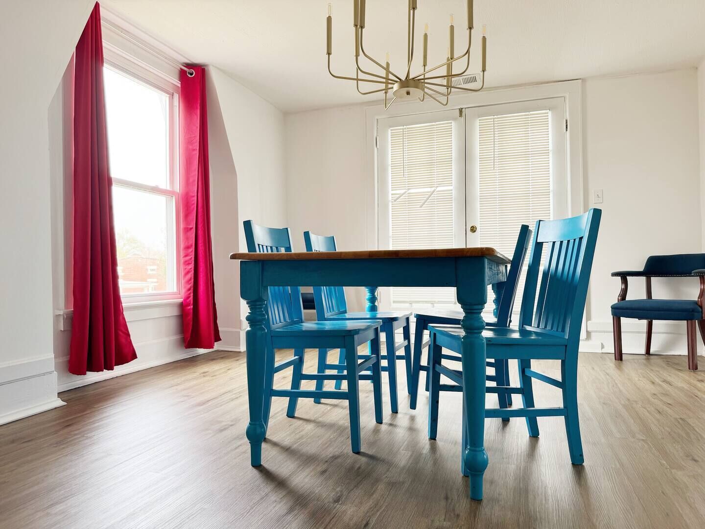 Un coin repas avec une table en bois et des chaises bleues, un lustre et une fenêtre ornée de rideaux rouges.