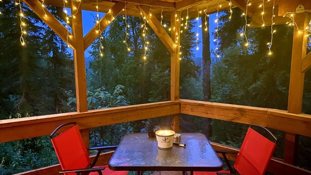 A covered outdoor seating area with a table and chairs, string lights, and a view of trees. - Bear Cabin-Gunning Cabin Rentals (Soldotna)