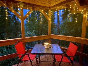 A covered outdoor seating area with a table and chairs, string lights, and a view of trees. - Bear Cabin-Gunning Cabin Rentals (Soldotna)