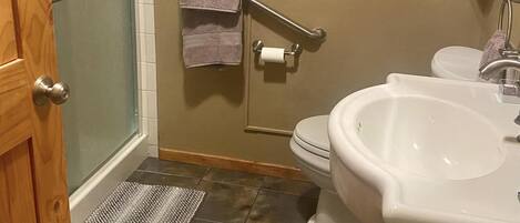 Une salle de bain équipée d’une douche, de toilettes et d’un lavabo. On y trouve des barres d’appui, un porte-serviettes et un tapis au sol.