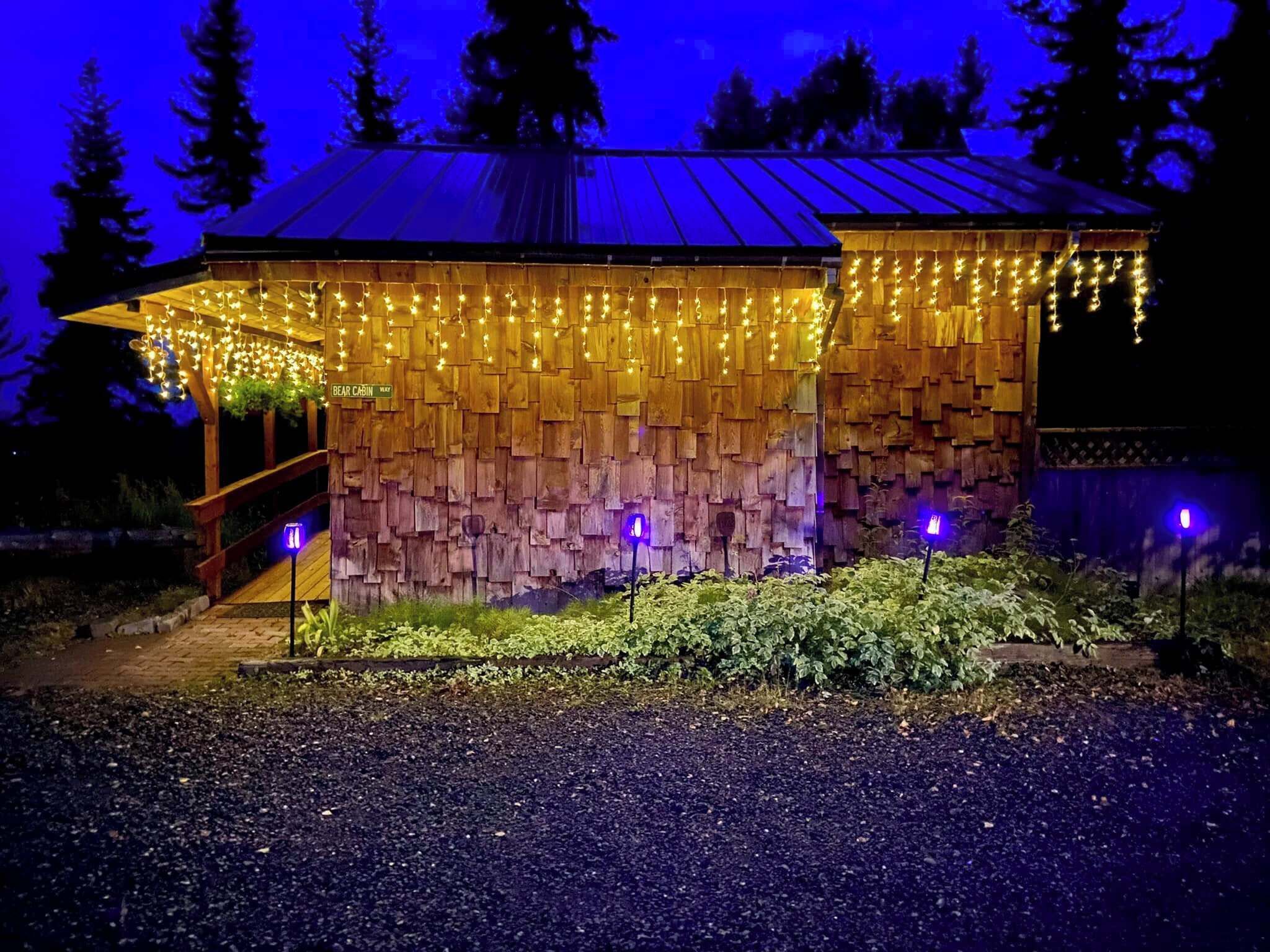 Una cabaña rústica con techo inclinado, paredes de madera y un porche decorado con luces colgantes.