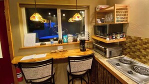 A kitchen with a wooden countertop, two chairs, a stove, a microwave, and a window with stained glass lamps. - Bear Cabin-Gunning Cabin Rentals (Soldotna)