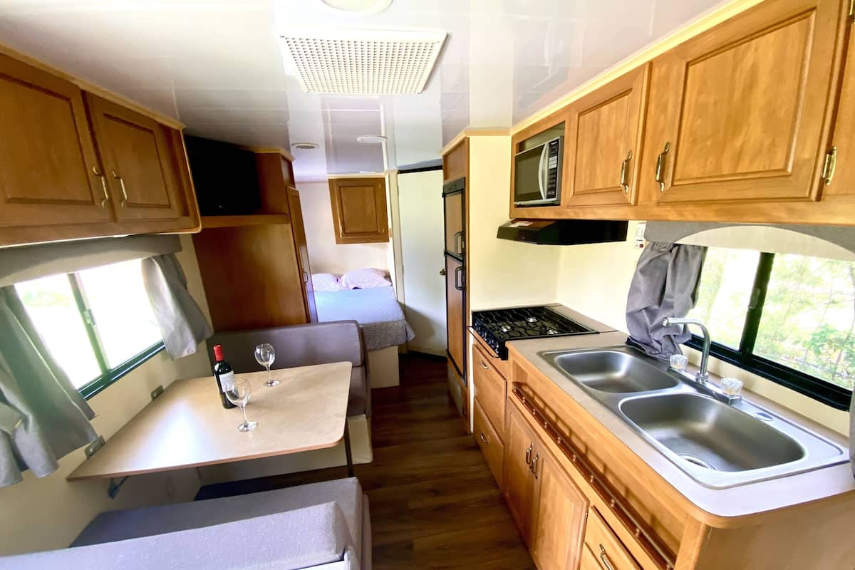 Une cuisine compacte pour camping-car, dotée de meubles en bois, d’une cuisinière, d’un évier et d’un petit coin repas avec une table et deux verres à vin.
