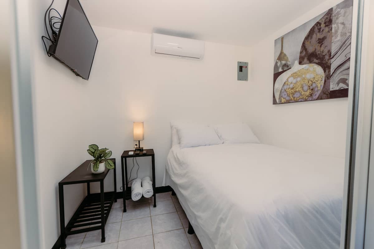 Une chambre d’hôtel avec un lit, un téléviseur fixé au mur, un appareil de climatisation, une table de chevet avec une lampe et un tableau au mur.
