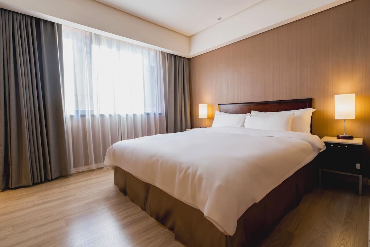 A hotel room with a large bed, two bedside lamps, a wooden headboard, and a view of the outdoors through sheer curtains.