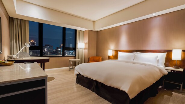 Habitación de hotel con una cama grande, un escritorio, una silla, una mesita de noche y vistas de la ciudad de noche.
