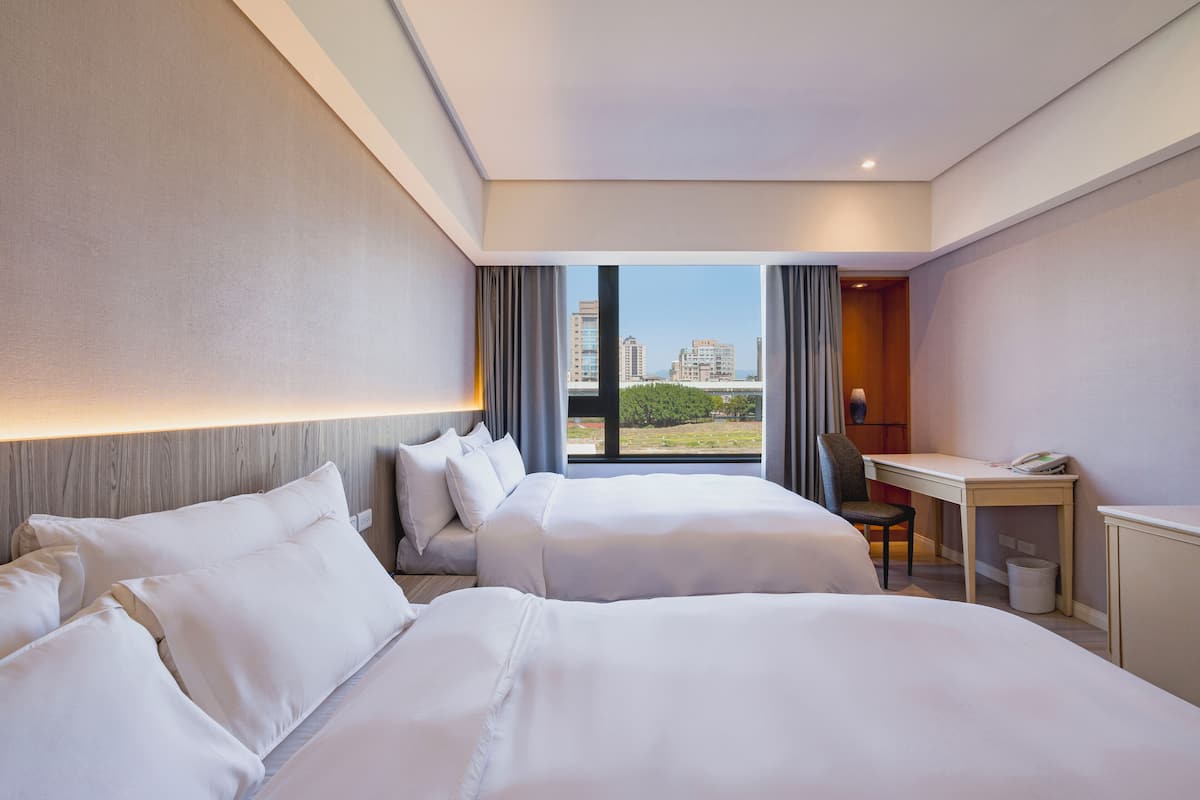 A hotel room with two beds, a desk, and a view of buildings.