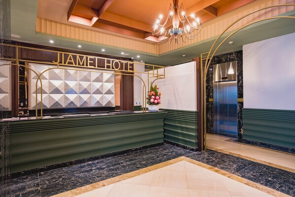 Uma área de recepção de hotel com design moderno, destacando um balcão de atendimento, um candelabro e uma parede decorativa com padrões geométricos.