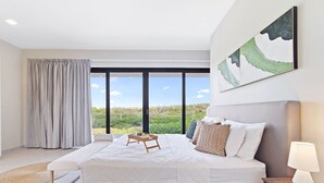 A modern bedroom with a large bed, a bedside table with a lamp, a view of greenery through large windows, and a painting on the wall. - 4BR Private Pool & Jacuzzi Boca Catalina Beach (Noord)