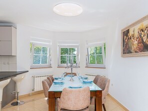 A dining room with a large table set for six, flanked by three windows with white frames.