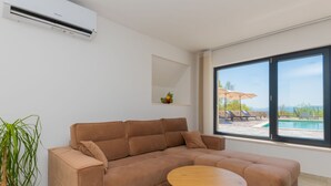 Ein modernes Wohnzimmer mit einem braunen Sofa, einem kleinen Holztisch, einer Topfpflanze und einem großen Fenster mit Blick auf einen Pool.