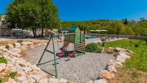 Ein Spielplatz mit Schaukeln und einer kleinen Spielhütte, umgeben von Kiesflächen und einem Pool mit Liegestühlen.
