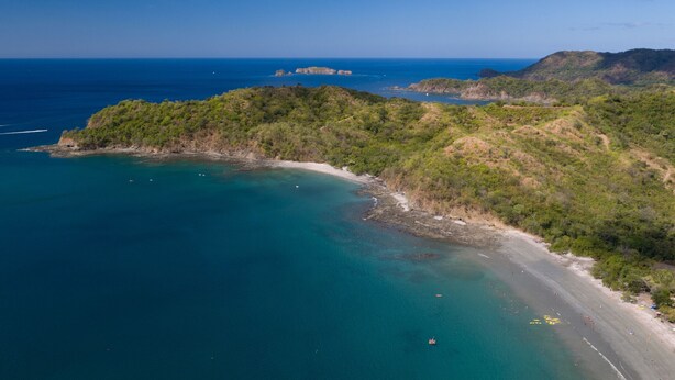 Vista aérea de una zona costera con playa, aguas azules cristalinas y abundante vegetación.