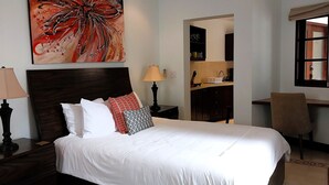 Una cama bien tendida con sábanas blancas y varios almohadones, un cabecero de madera oscura, una lámpara de noche y un cuadro abstracto de gran tamaño sobre la cama.