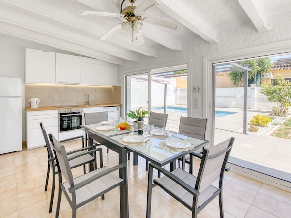 Una cocina moderna con armarios blancos, un comedor con mesa de vidrio y vistas a una piscina.