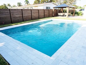 A rectangular swimming pool with a tiled deck, surrounded by a wooden fence and a covered patio area. - Heated Pool House Near Downtown West Palm Beach, Beaches, and Bars. (West Palm Beach)