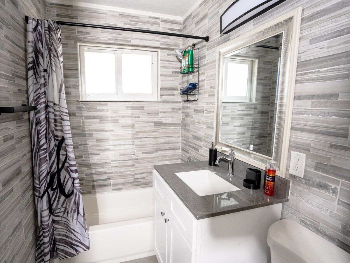 Une salle de bain moderne avec un mur recouvert de carreaux gris et blancs, un grand miroir au-dessus du lavabo et un rideau de douche orné d’un motif de feuilles.