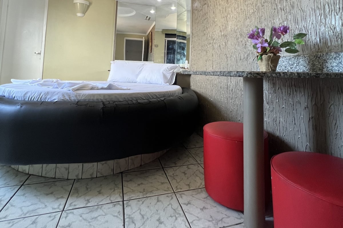A modern hotel room with a bed, a red ottoman, a small table with a vase of flowers, and a textured wall.