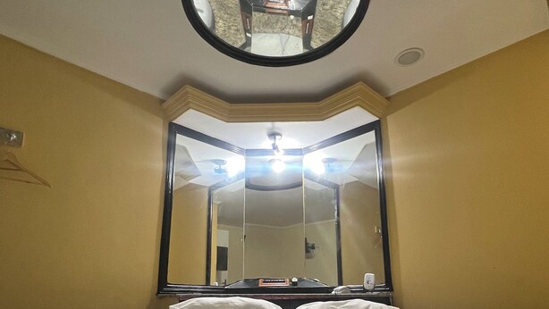 Habitación de hotel con un espejo redondo sobre la cama, un ventilador de techo y un espejo grande que refleja la habitación.