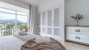 Une chambre avec un grand lit, une vue sur l’extérieur, une commode blanche et un vase rempli de fleurs.