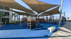 Un área de juegos al aire libre con tobogán, estructuras para escalar y asientos a la sombra.
