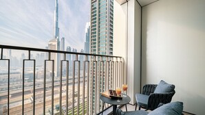 A balcony with a view of the Burj Khalifa and other buildings, two grey armchairs, a small table with a drink, and a book.