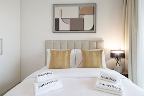 A hotel room with a bed, pillows, a headboard, a nightstand with a lamp, and a framed artwork on the wall.