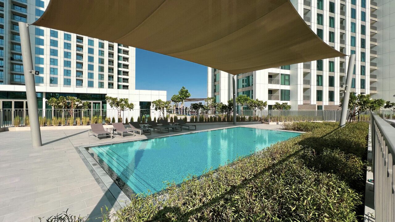 A swimming pool with lounge chairs, surrounded by a fence and greenery, with modern buildings in the background.