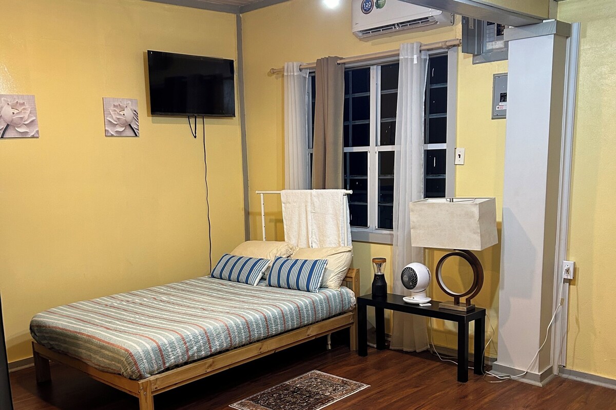 A bedroom with a wooden bed, a small table with a lamp, a wall-mounted TV, and a window with curtains.