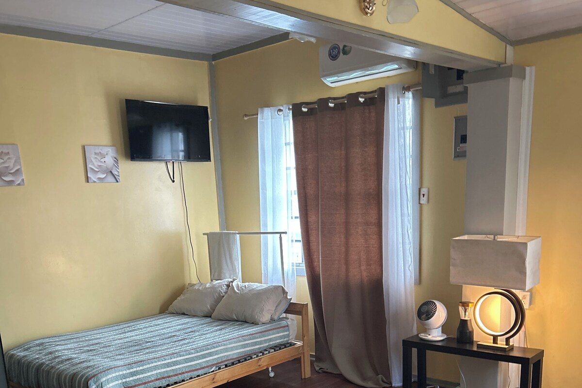 A small, cozy room with a single bed, a bedside table with a lamp, a wall-mounted TV, and a window with curtains.