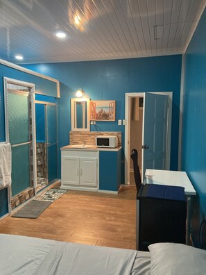 A room with blue walls, a wooden floor, a white cabinet, a microwave, a sink, a door, and a bed.
