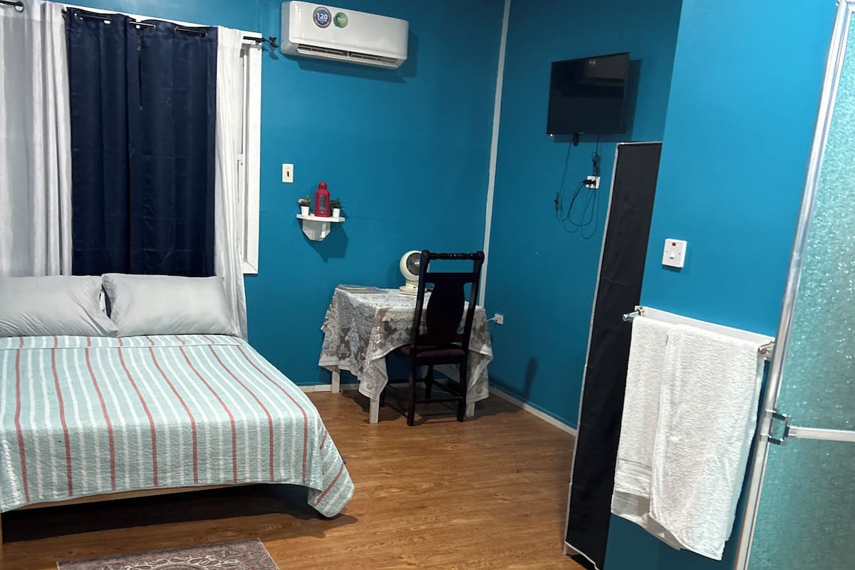A room with a bed, a small table with a chair, an air conditioner, a refrigerator, and a shower area.