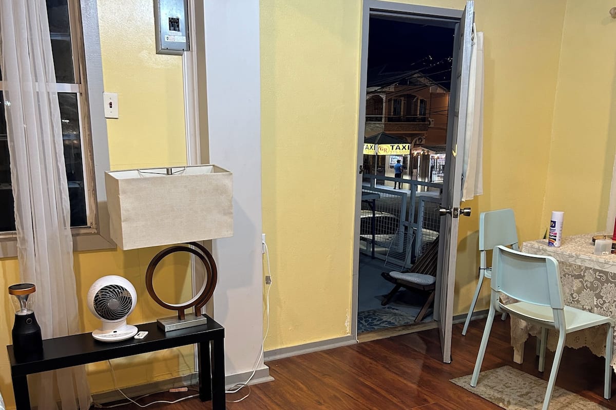 A room with a wooden floor, a dining table with chairs, a small table with a lamp, and a window with curtains.
