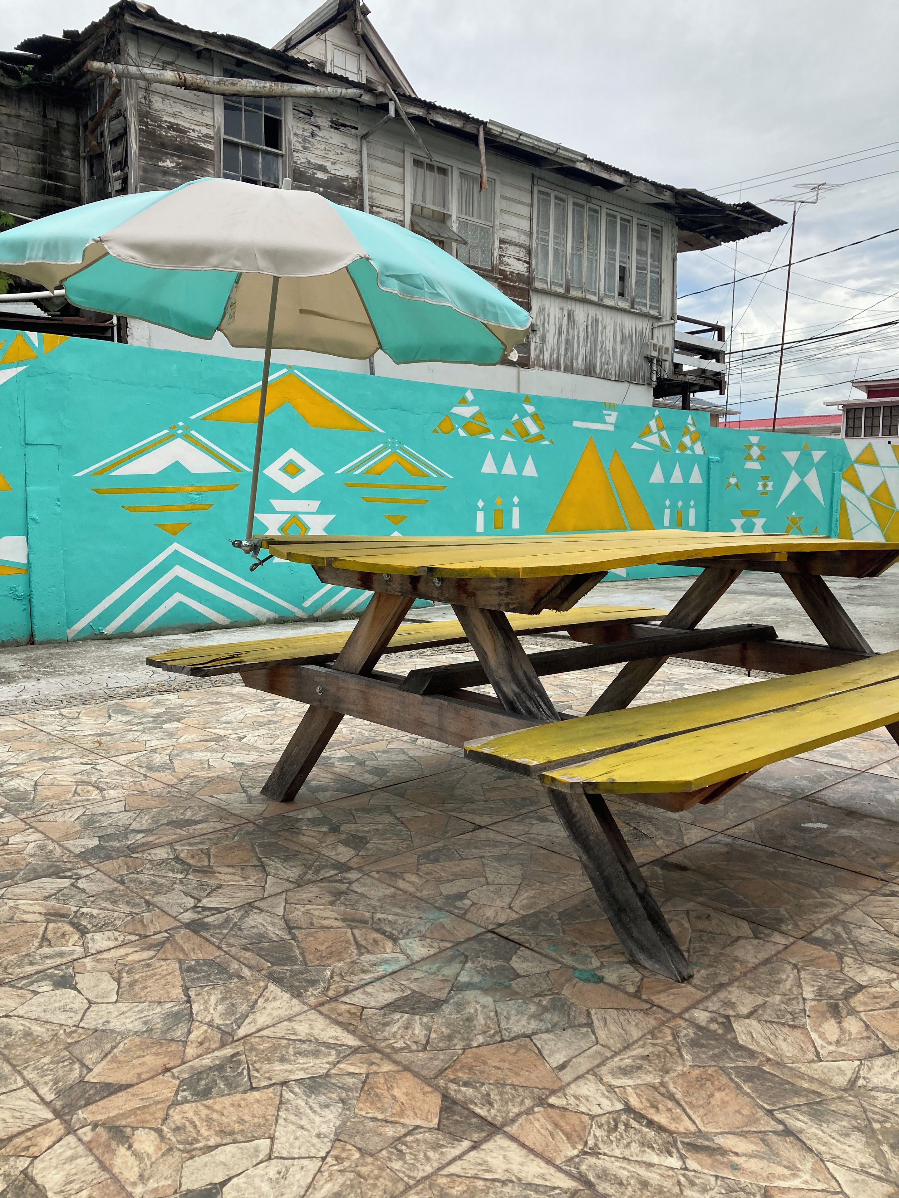 A picnic table with a patterned umbrella in front of a building with geometric wall art.